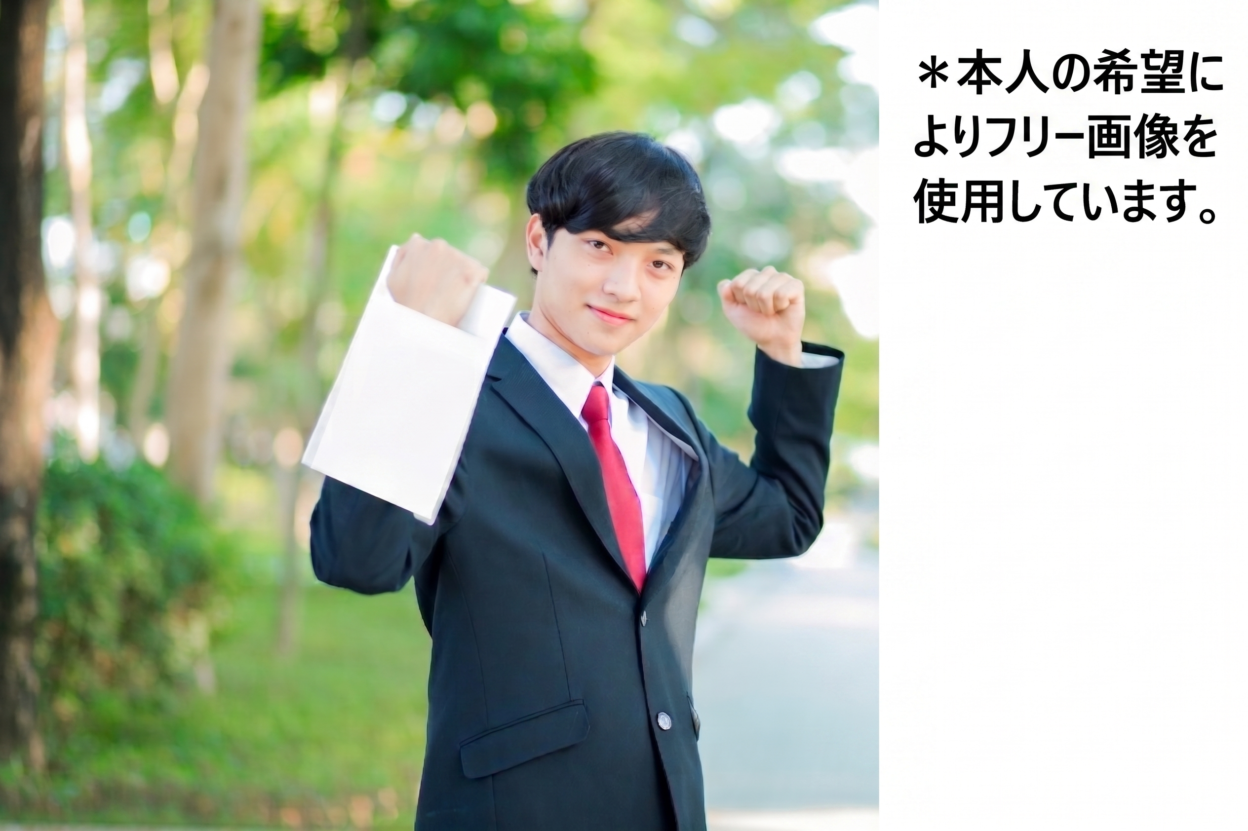 Outdoor portrait of a young man in a dark suit and red tie, raising his fists in a celebratory pose while holding a white shopping bag, with a blurred green park background (and Japanese disclaimer text on the white panel to the right).