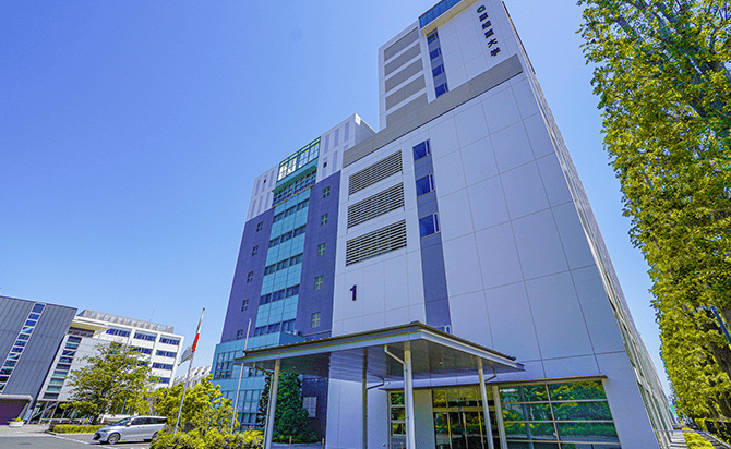 Modern hospital building with a glass entrance and blue-gray exterior under a clear blue sky while trees surround the scene.