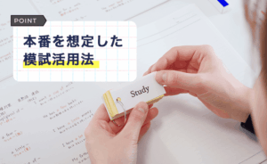 Close-up of hands holding a small 'Study' tag and a card reading '本番を想定した 模試活用法', labeled as a study tip section on a desk.